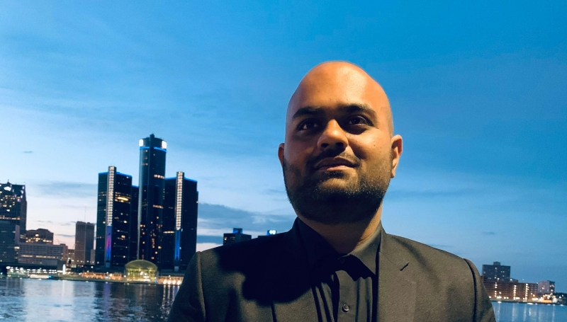 Kushal Patel standing in front of the Detroit river with the GM Renaissance Center in the background.